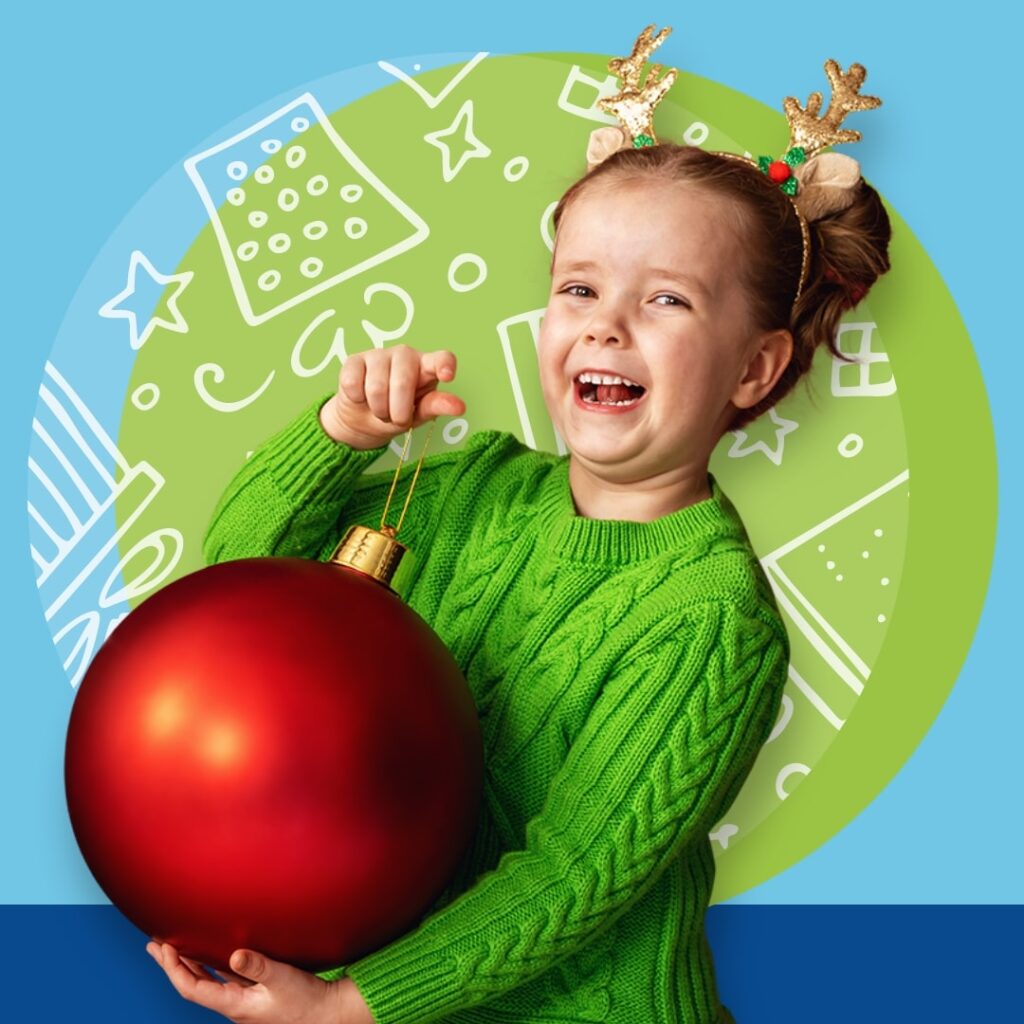 Little girl wearing reindeer antlers holding large ornament, laughing