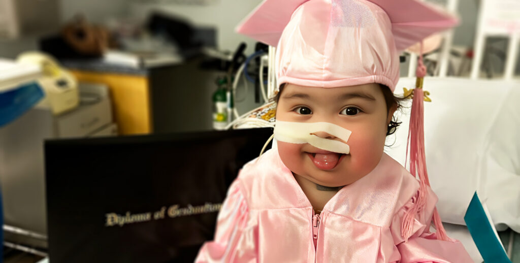 toddler girl with surgical tape cover feeding tube on her face, wearing a pink graduation cap and gown