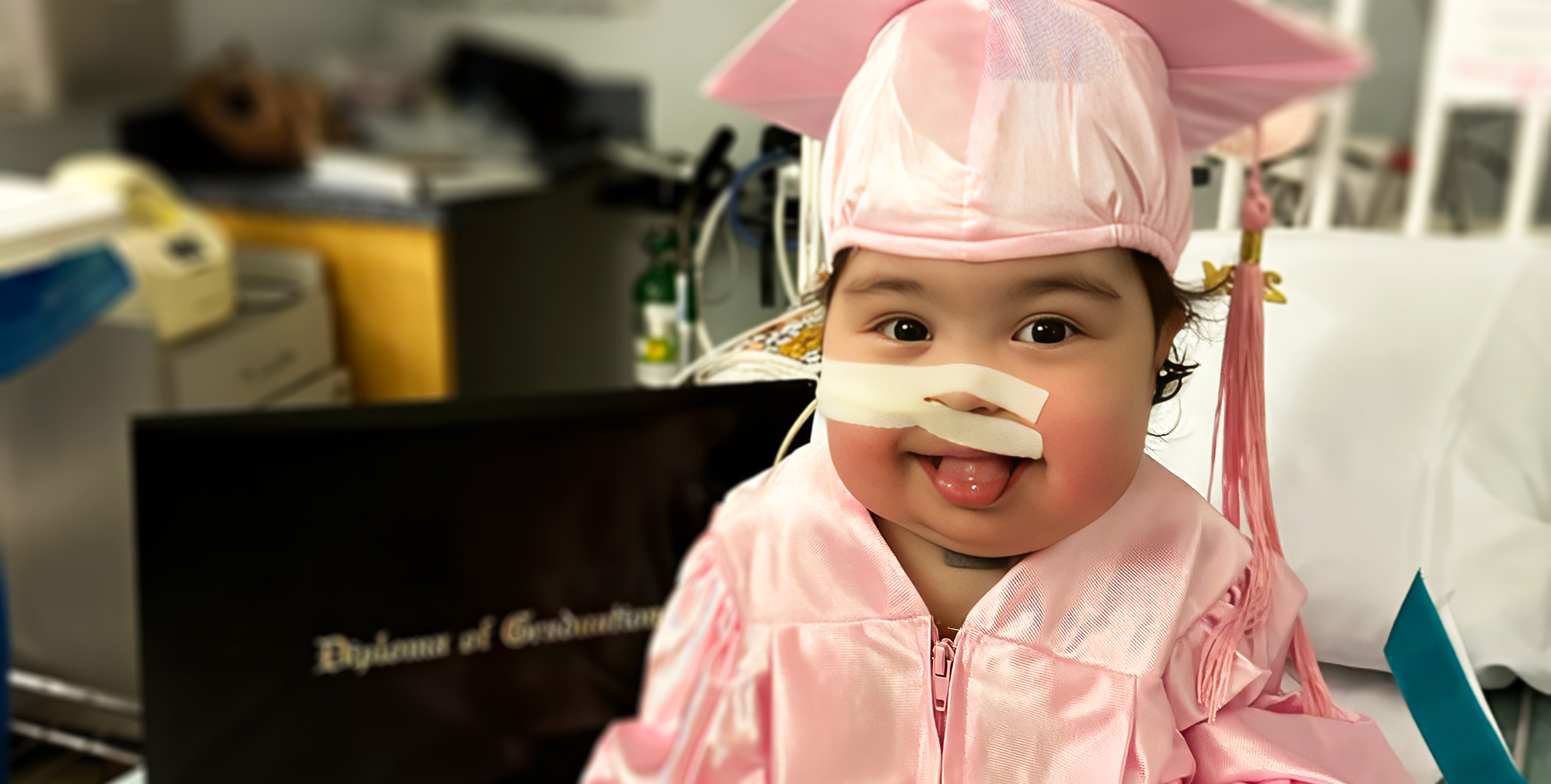 toddler girl with surgical tape cover feeding tube on her face, wearing a pink graduation cap and gown