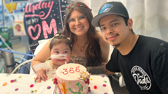 toddler girl with surgical tape covering feeding tube on her face, with mom, dad and celebration cake.