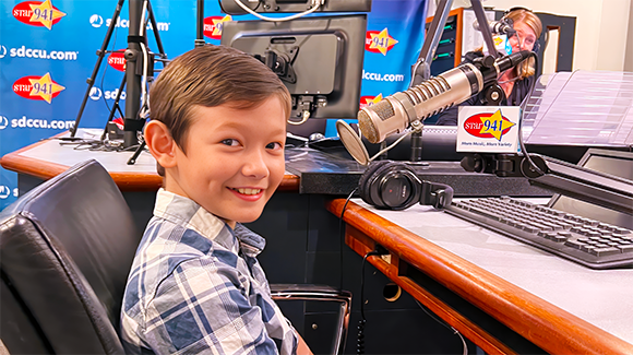boy with brown hair, wearing blue and white shirt and smiling, waiting to be interviewed at fundraising event.