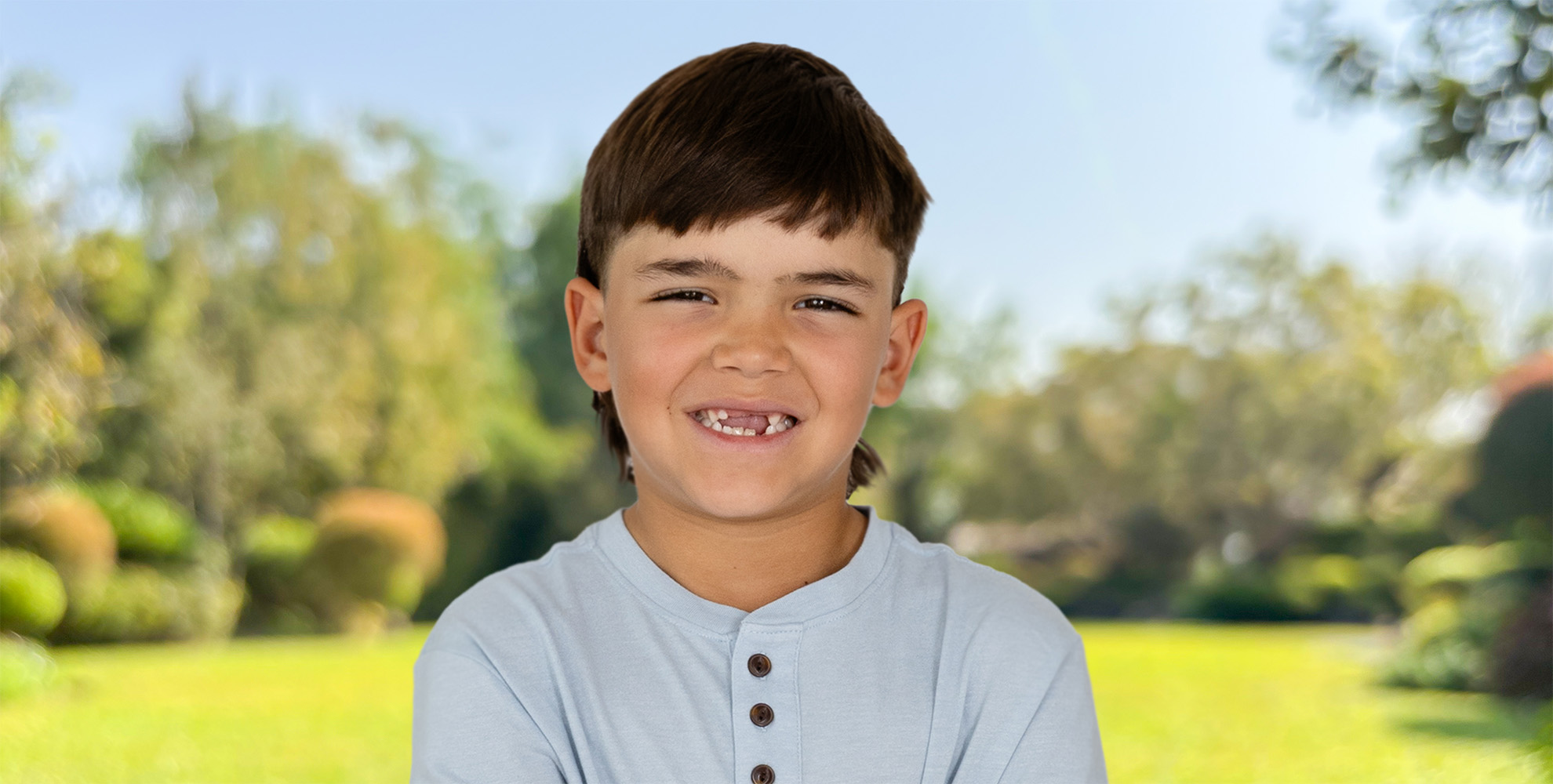 Boy with brown hair and missing front teeth smiling in outdoor setting