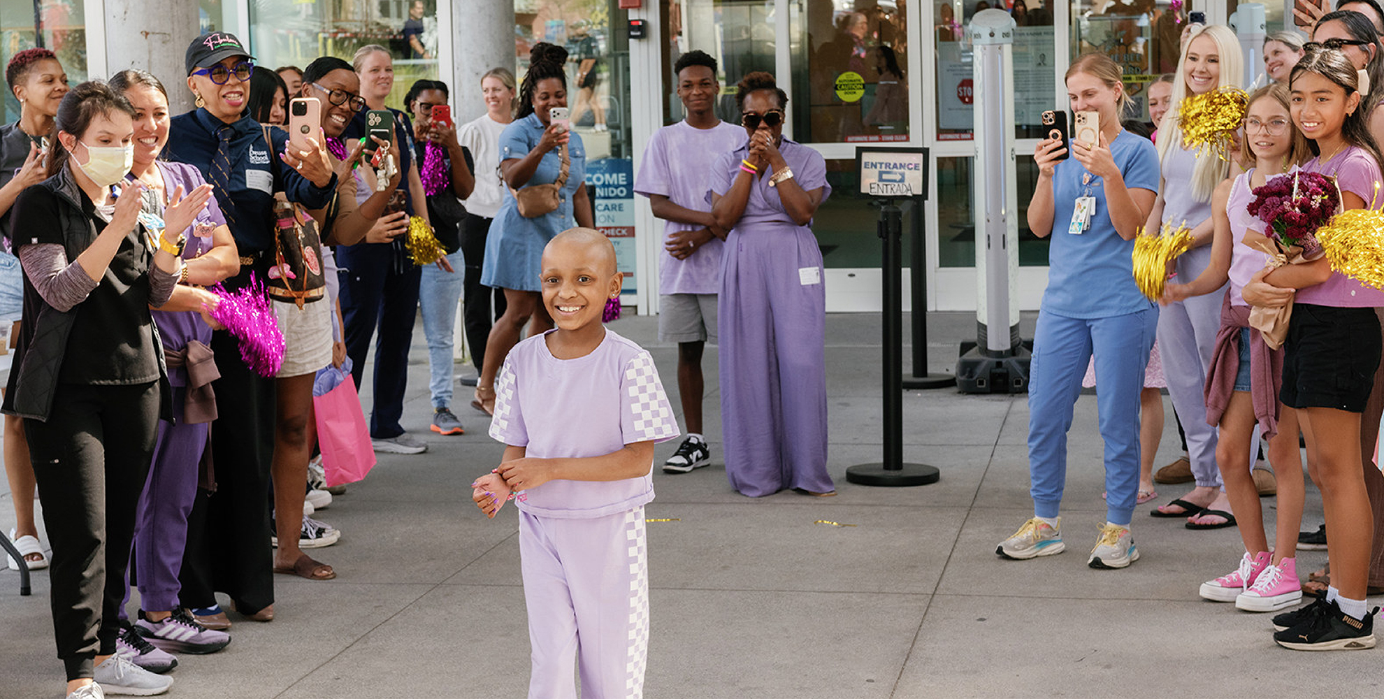 Girl without hair wearing purple outfit walking out of hospital with crowd cheering around her