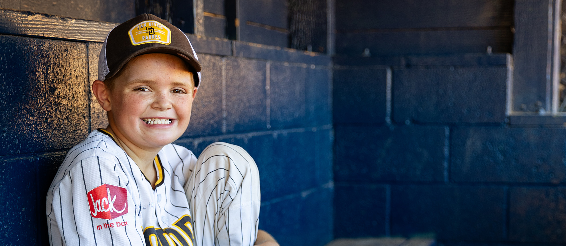 Boy wearing baseball hat and uniform sitting in dugout